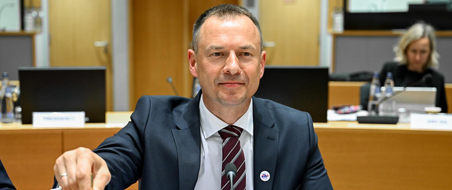 Minister Piotr Borys siedzi za stołem. Przed nim na blacie leżą dokumenty. W prawej ręce minister trzyma dzwonek.