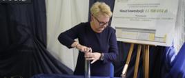 Kobieta w granatowej sukience stoi nad nakrytym niebieskim materiałem stołem, trzyma błyszczący metalowy cylinder, za nią tablica z napisem Koszt inwestycji 11 708 010 zł.