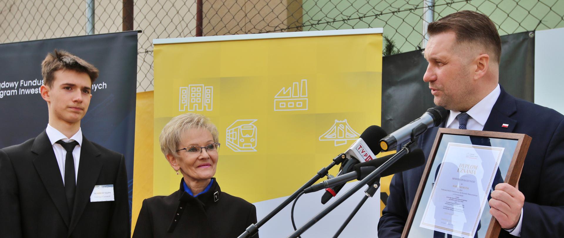 Minister Czarnek stoi za drewnianą mównicą i pokazuje trzymany w ręku dokument w ramkach, przed nim kilka mikrofonów, obok stoi ubrana na czarno kobieta w okularach i młody człowiek w garniturze, za nimi ogrodzenie z siatki.