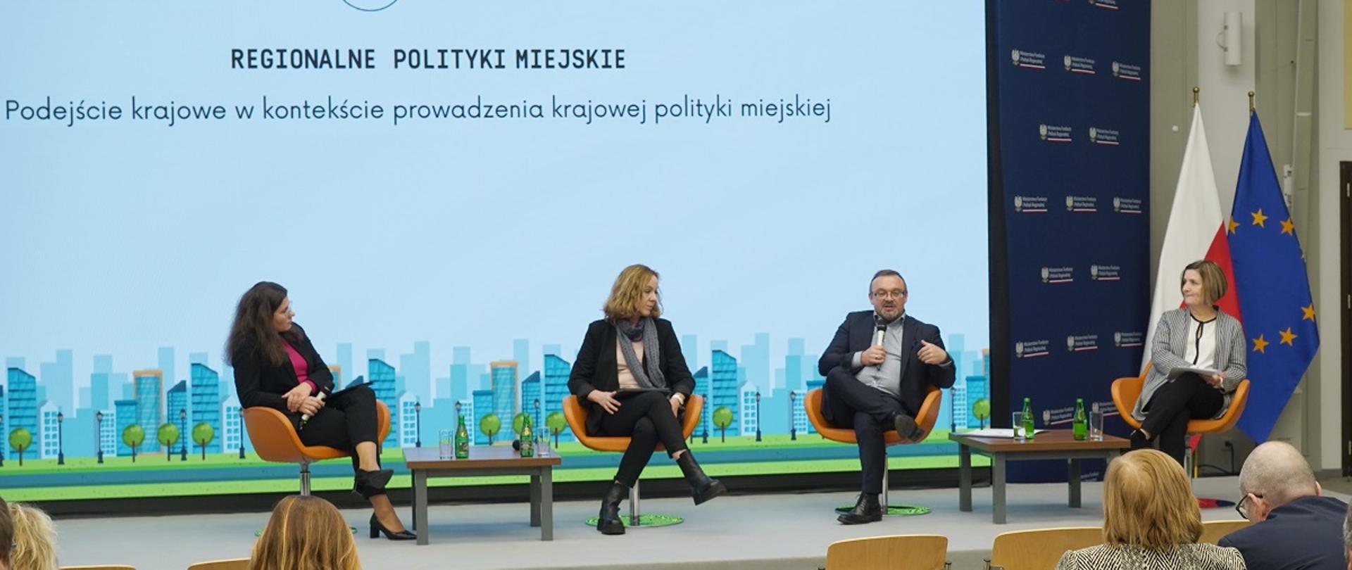Obrady V Krajowego Forum Miejskiego (KFM 2023), paneliści spotkania siedzą w fotelach i prowadzą dyskusję, za nimi ekran z grafiką informującą o obradach KFM