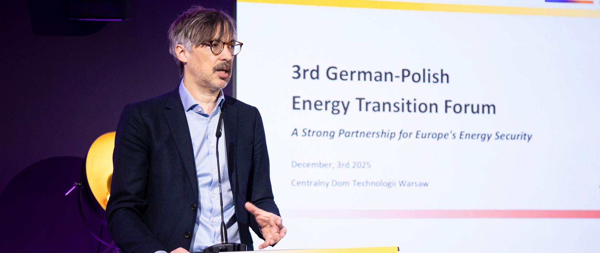 Wiceminister klimatu i środowiska Krzysztof Bolesta na III Polsko-Niemieckim Forum Transformacji Energetycznej. Wiceminister stoi za mównicą. Za nim prezentacja z napisem Trzecie Polsko-Niemieckie Forum Transformacji Energetycznej 