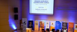sala konferencyjna, z rzędami krzeseł dla publiczności. Na scenie znajduje się mównica, za którą stoi kobieta. W tle widać banery w różnych kolorach z napisami „BELNY SZKOŁNY” i logo Uniwersytetu Gdańskiego. Na dużym ekranie nad sceną widnieje napis „KONGRES SZKOLNYCH KASZËBSCZÉGÒ JÃZËKA Gdańsk, 13 sãwnika 2025 roku” oraz nazwisko prelegentki „BARBARA NOWACKA Ministrõ Nãrõdny Edukacje”.