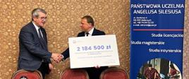 Za drewnianym stołem stoi minister Murdzek i mężczyzna w garniturze, podają sobie ręce, mężczyzna trzyma duży symboliczny czek z napisem 2 184 500 zł.