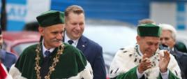 Dwóch mężczyzn w zielonych ceremonialnych strojach idzie ulicą, obaj się śmieją, jeden klaszcze, za nimi minister Czarnek.
