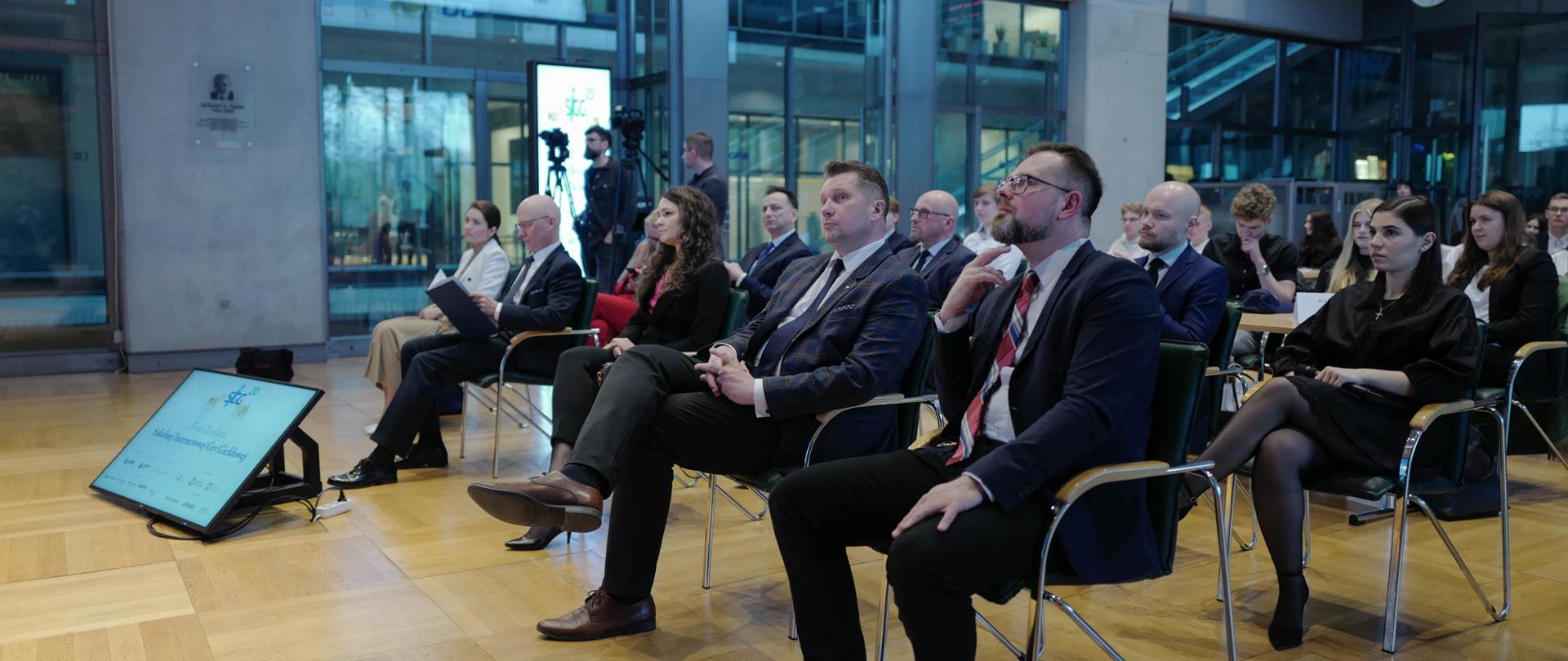 Grupa osób siedzących w kilku rządach krzeseł w dużej, przestronnej sali z dużymi oknami. W pierwszym rzędzie minister Czarnek trzyma "nogę na nogę". Na drewnianej podłodze, przed pierwszym rzędem leży ekran, na którym się coś wyświetla. 