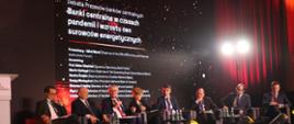 Kobieta i mężczyźni siedzą na scenie, uczestniczą w debacie, za nimi napis Banki Centralne w czasach pandemii wzrostu cen surowców energetycznych 