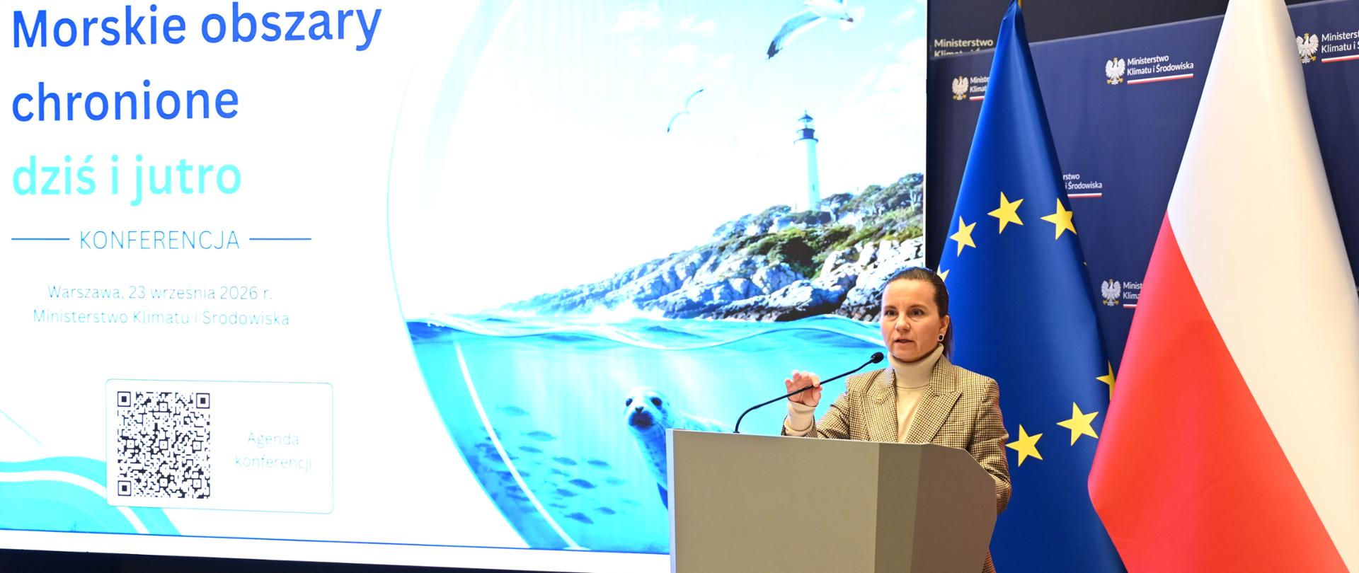 Urszula Zielińska, sekretarz stanu w Ministerstwie Klimatu i Środowiska stoi przy mównicy i przemawia podczas konferencji. Za mównicą znajdują się flagi Polski i Unii Europejskiej. Na dużym ekranie w tle wyświetlono ilustrację podwodnego środowiska morskiego z foką i ławicą ryb. Zdjęcie wykonano wewnątrz sali konferencyjnej i nawiązuje do tematyki ochrony przyrody oraz środowiska morskiego.