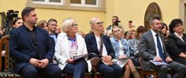 Grupa ludzi siedzi na krzesłach, za nimi żółta ściana z oknem. Wszyscy patrzą w lewo.