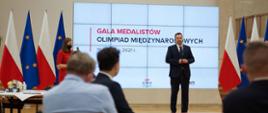 Minister Edukacji i Nauki Przemysław Czarnek przemawia na tle wyświetlonego napisu "Gala Medalistów Olimpiad Międzynarodowych. W tle flagi Polski i UE