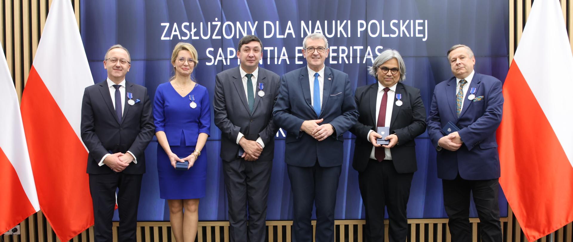 Pod wbudowanym w ścianę wielkie ekranem z napisem Ceremonia wręczenia medali „Zasłużony dla Nauki Polskiej Sapientia et Veritas” stoi pięciu mężczyzn w garniturach i kobieta w niebieskim ubraniu, po bokach polskie flagi.