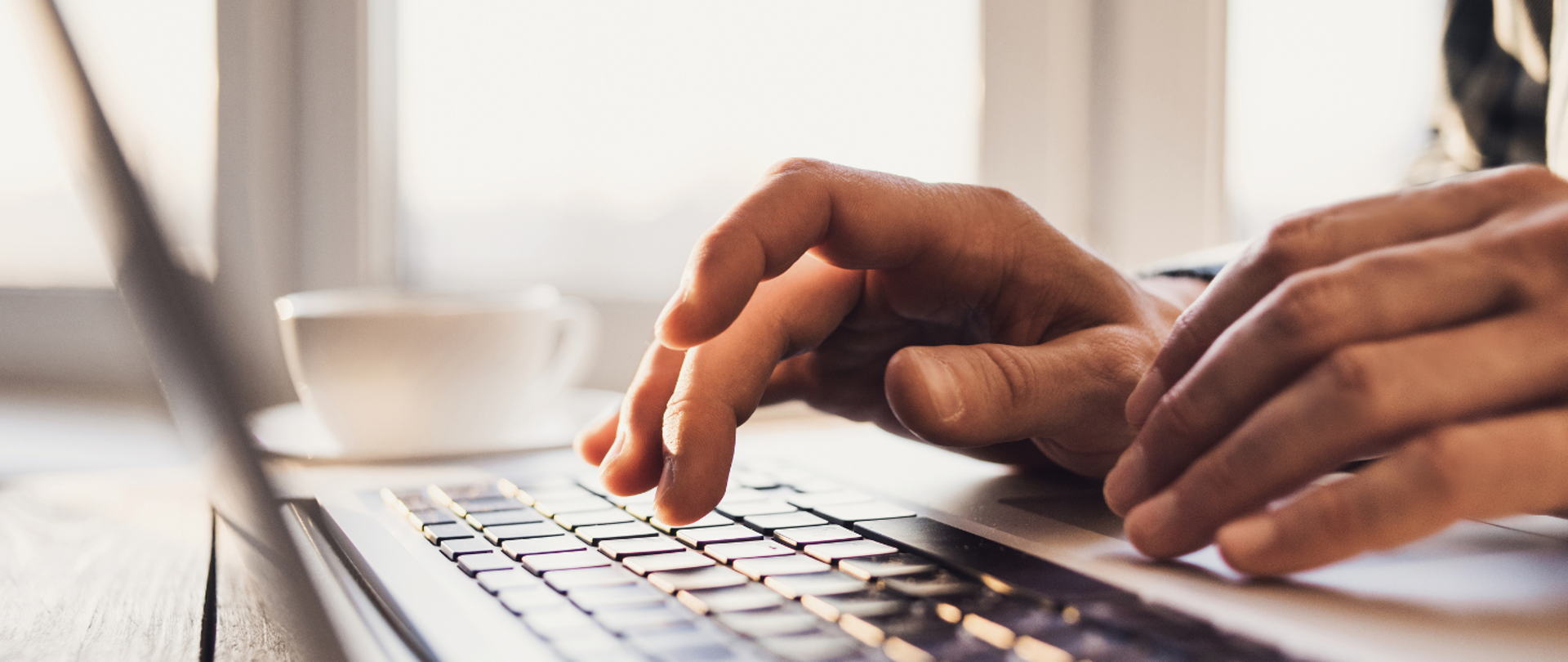 Na grafice widać przybliżoną klawiaturę laptopa oraz męskie dłonie unoszące się nad klawiaturą. W tle widać rozmytą białą filiżankę.