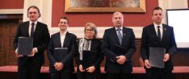 Pięć osób stoi przed stołem, w rękach trzymają czarne teczki, za nimi na czerwonej ścianie obraz.