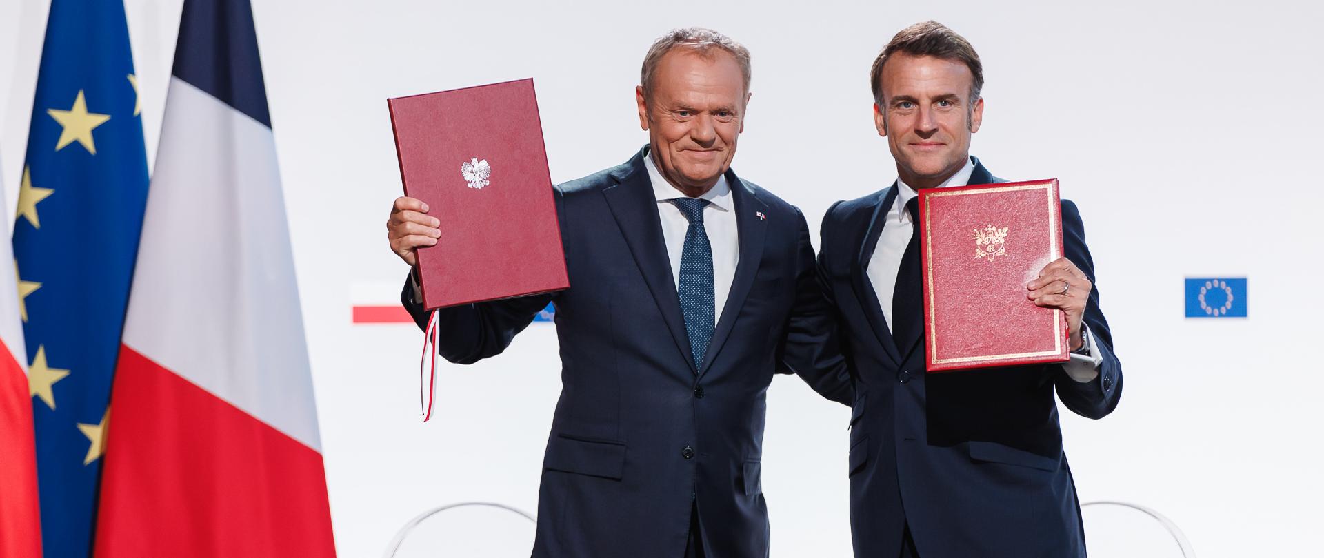 Premier Donald Tusk i Prezydent Francji Emmanuel Macron po podpisaniu traktatu polsko-francuskiego prezentują teczki z podpisanym dokumentem. W tle flagi Polski, Francji i Unii Europejskiej.