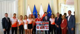 Minister sportu i turystyki Kamil Bortniczuk stoi wraz z kajakarzami, medalistami ostatnich mistrzostw świata w kajakarstwie. Wraz z ministrem stoi jedenaście osób, na tle flag Polski i Unii Europejskiej. Jeden z zawodników trzyma razem z ministrem prezent, pamiątkowe zdjęcia, które zawodnicy przekazali ministrowi.
