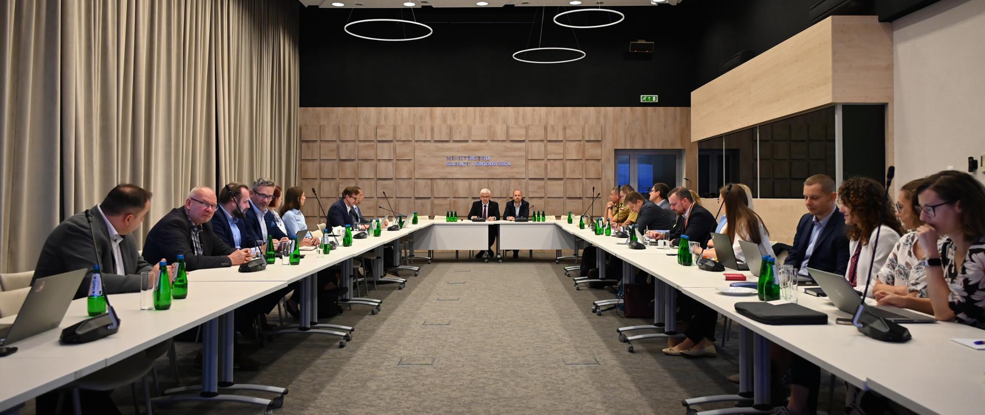 Wiceminister klimatu i środowiska Ireneusz Zyska oraz zastępca dyrektora Departamentu Elektromobilności i Gospodarki Wodorowej Marek Popiołek wraz z uczestnikami podczas spotkania Komitetu Sterującego ds. Elektromobilności siedzą przy stole w sali konferencyjnej.