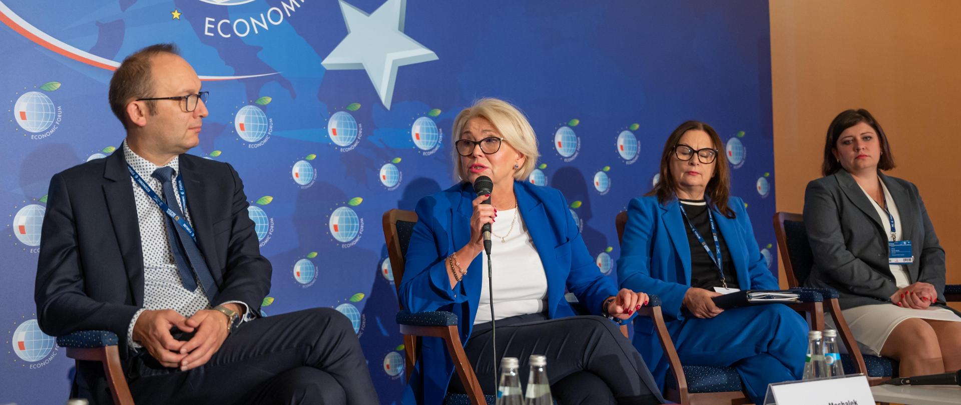 Wiceminister Machałek i kilka innych osób siedzą na krzesłach, za nimi niebieska ściana, minister Machałek mówi do trzymanego w ręku mikrofonu.