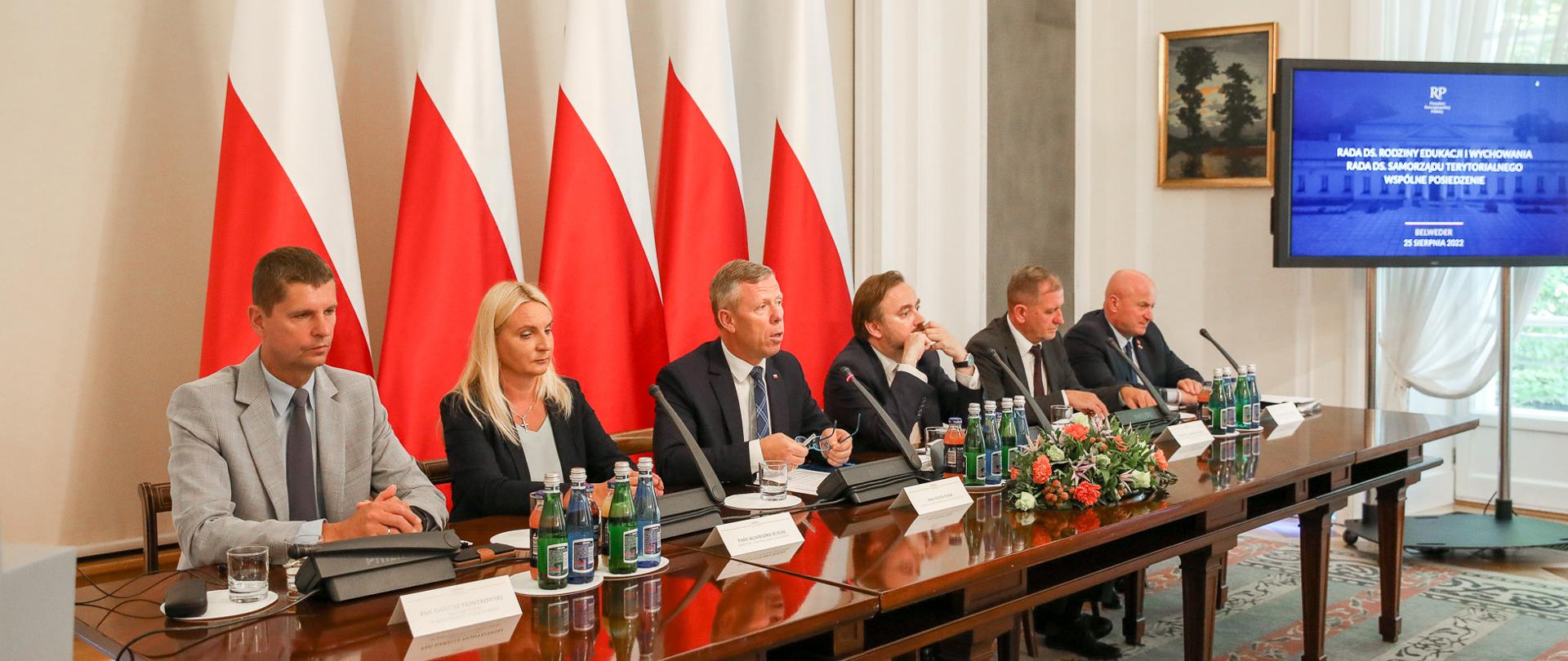 Minister Piontkowski siedzący przy stole w towarzystwie czterech mężczyzn i jednej kobiety. Za nimi pięć flag Polski