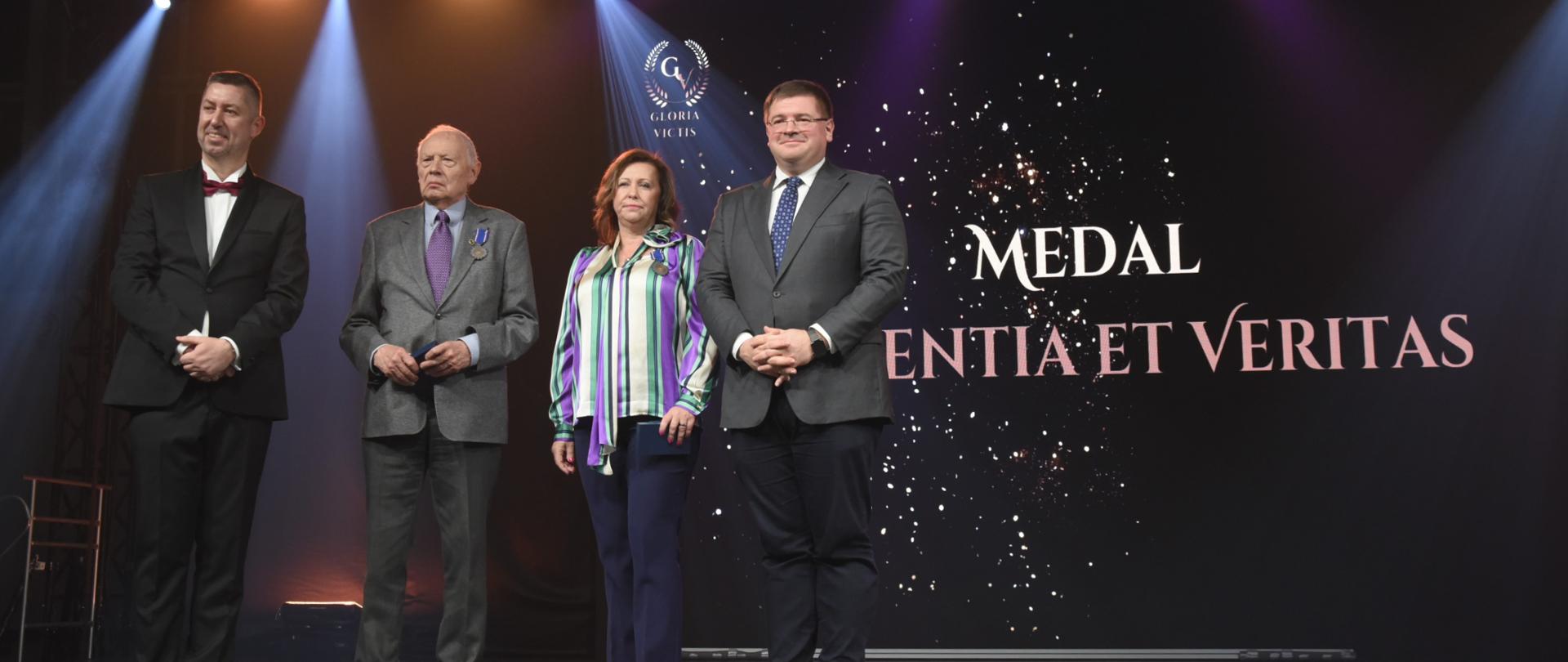 Zdjęcie przedstawia stojących na scenie trzech mężczyzn oraz jedną kobietę, w tle ciemny ekran z napisami oraz reflektory. Jeden mężczyzna i kobieta mają przypięte medale.