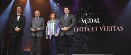 Zdjęcie przedstawia stojących na scenie trzech mężczyzn oraz jedną kobietę, w tle ciemny ekran z napisami oraz reflektory. Jeden mężczyzna i kobieta mają przypięte medale.