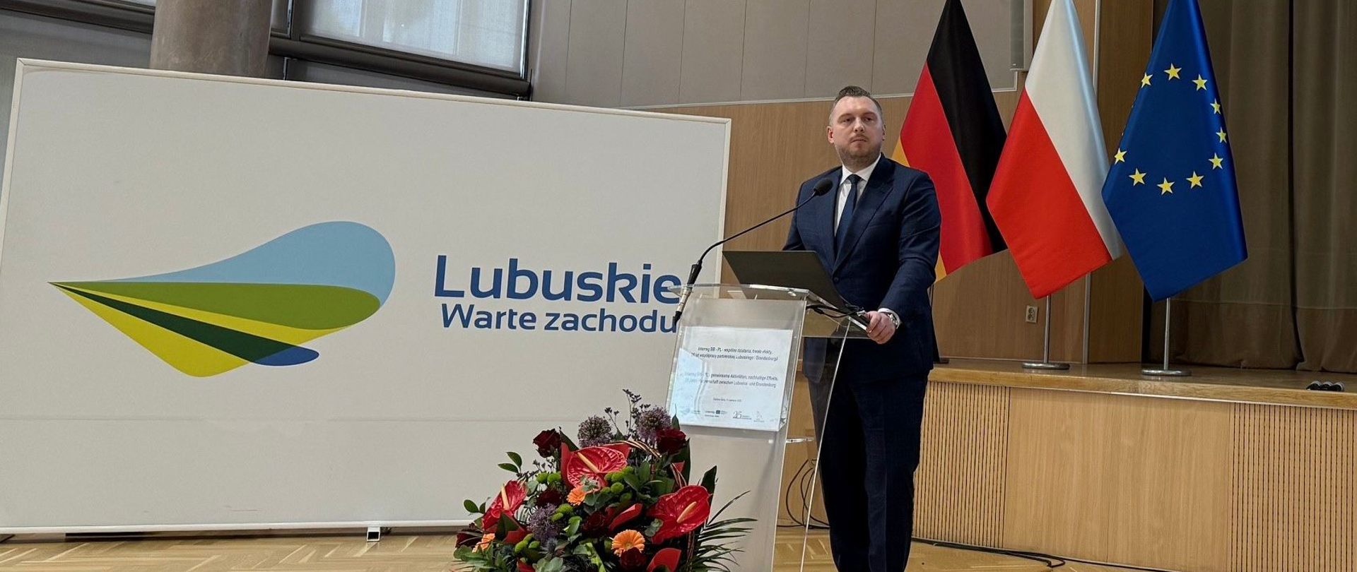 W sali konferencyjnej wiceminister Konrad Wojnarowski stoi w mównicy. Przed nim kosz z kwiatami. Za nim flagi Polski, Niemiec i UE.