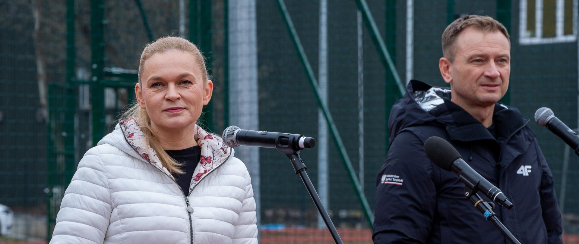 Minister Barbara Nowacka i minister sportu Sławomir Nitras stoją przy mikrofonach. W tle widać sportową bieżnię.
