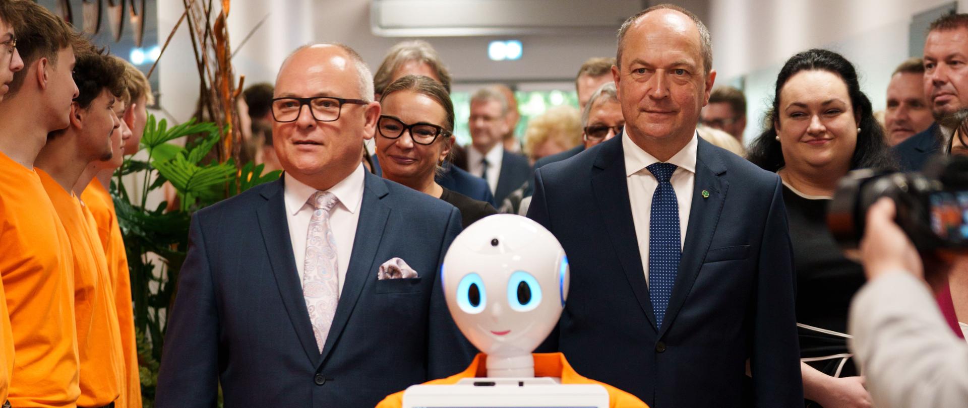 Duże wydarzenie wewnątrz budynku, gdzie w centrum zdjęcia dwóch mężczyzn w garniturach stoi obok humanoidalnego robota w pomarańczowej koszulce z napisem "BCU". Mężczyzna po lewej stronie, w okularach, patrzy wprost, uśmiechając się. Mężczyzna po prawej stronie, bez okularów, patrzy prosto przed siebie. Po lewej i prawej stronie widoczne są inne osoby, w tym grupa młodych ludzi w pomarańczowych koszulkach. W tle znajduje się korytarz.