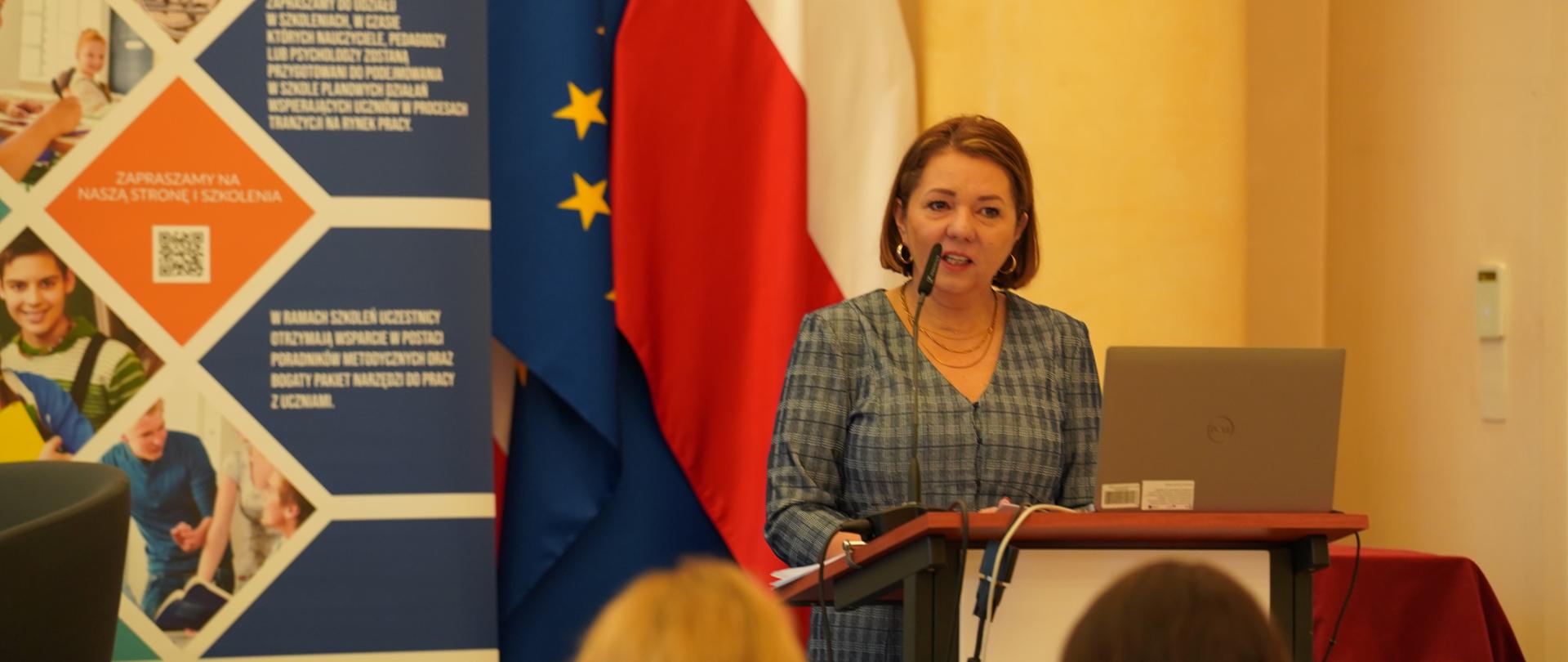 Kobieta w szarej sukience w drobną kratkę stoi za mównicą, przemawiając do mikrofonu. W tle widoczne są flagi Unii Europejskiej i Polski oraz pionowy, kolorowy baner promujący projekt "RYNEK PRACY – MOST DO PRZYSZŁOŚCI". Uczestnicy konferencji widoczni są na pierwszym planie od tyłu.