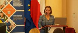 Kobieta w szarej sukience w drobną kratkę stoi za mównicą, przemawiając do mikrofonu. W tle widoczne są flagi Unii Europejskiej i Polski oraz pionowy, kolorowy baner promujący projekt "RYNEK PRACY – MOST DO PRZYSZŁOŚCI". Uczestnicy konferencji widoczni są na pierwszym planie od tyłu.