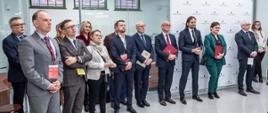 Duża grupa ludzi stoi w rzędzie, trzymają czerwone teczki z godłem, za nimi biała ściana.