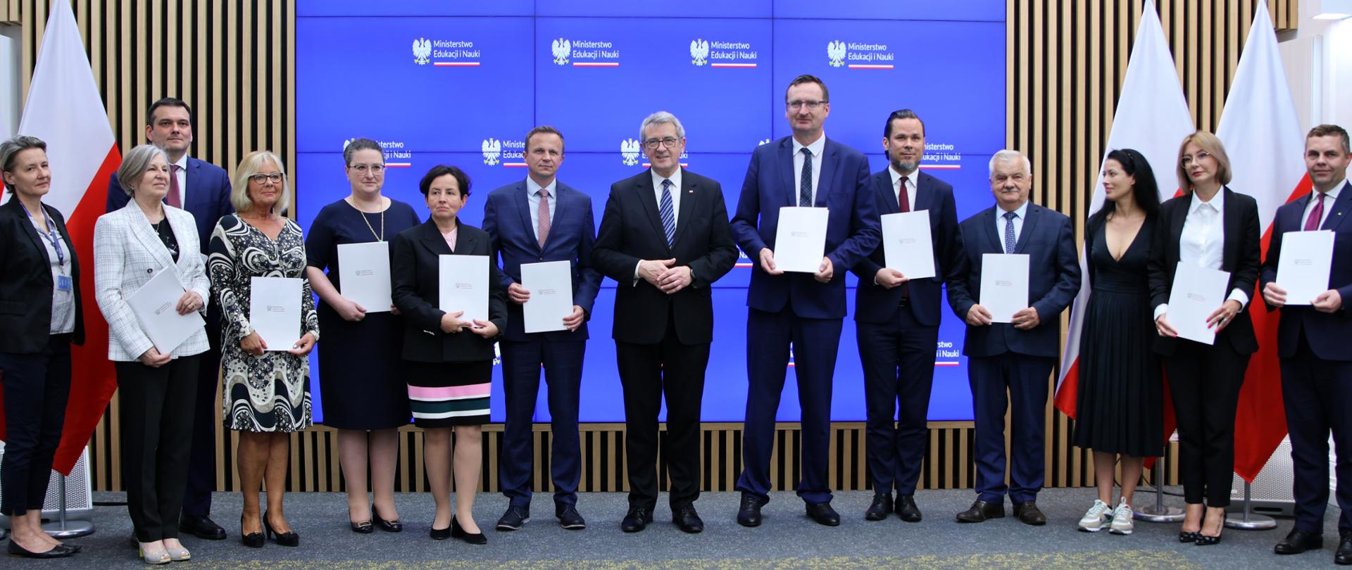 Grupa osób z białymi teczkami w rękach. W tle błękitny ekran z napisem Ministerstwo Edukacji i Nauki i orłem