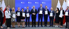 Grupa osób z białymi teczkami w rękach. W tle błękitny ekran z napisem Ministerstwo Edukacji i Nauki i orłem