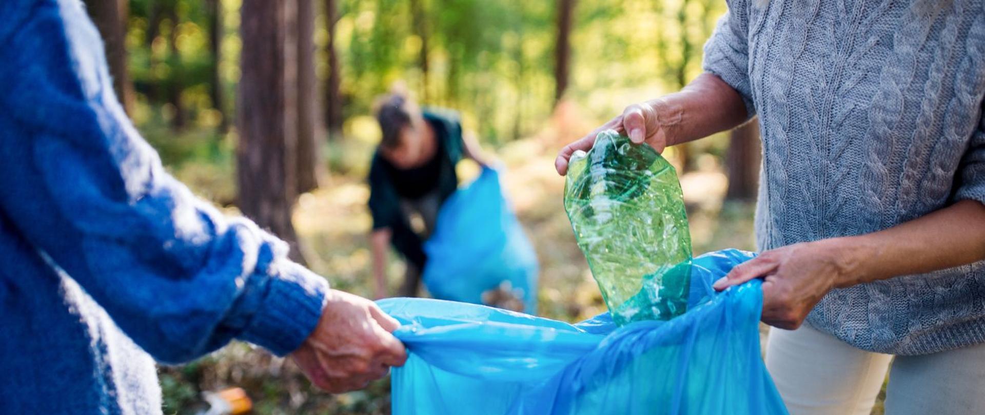 Na zdjęciu widoczni są ludzie, którzy sprzątają las. Zbierają plastikowe śmieci oraz plastikowe butelki do worków. Na pierwszym planie widoczne są dwie osoby, które trzymają niebieski worek i wrzucają do niego zużytą plastikową butelkę w zielonym kolorze.