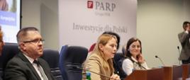 Minister funduszy i polityki regionalnej Katarzyna Pełczyńska-Nałęcz siedzi za stołem prezydialnym, po jej prawej stronie siedzi dyrektor Departamentu Programów Wsparcia Innowacji i Rozwoju w MFiPR Grzegorz Orawiec, zaś po lewej stronie siedzi Wojewoda Wielkopolski Agata Sobczyk