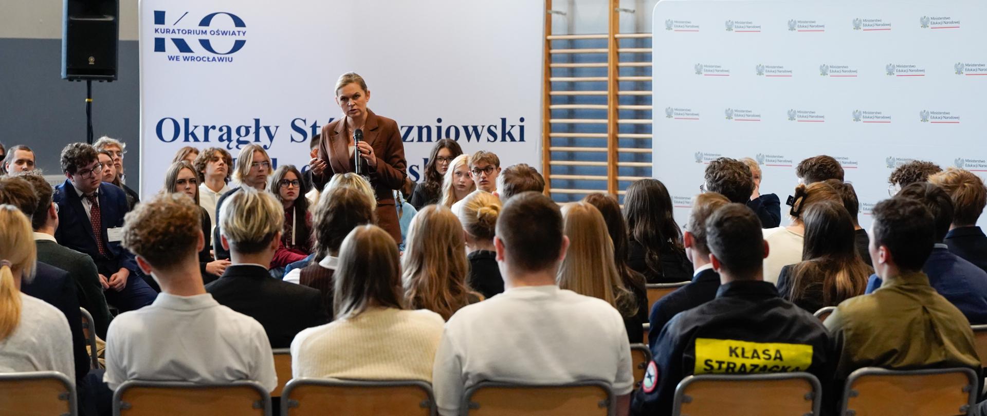 Minister Edukacji Barbara Nowacka stoi na tle ścianki z napisem "Okrągły Stół Uczniowski" i logotypem Kuratorium Oświaty we Wrocławiu, przemawiając do mikrofonu. Wzrok Minister Edukacji skierowany jest na uczniów, którzy siedzą wokół niej.