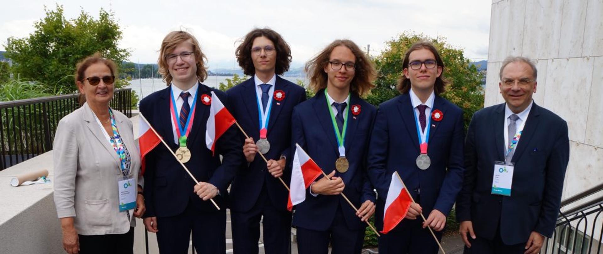 Czterech ubranych w garnitury laureatów trzymających małe polskie flagi stoi na tarasie, obok nich kobieta w białej marynarce i mężczyzna w garniturze.