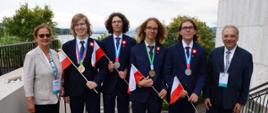 Czterech ubranych w garnitury laureatów trzymających małe polskie flagi stoi na tarasie, obok nich kobieta w białej marynarce i mężczyzna w garniturze.