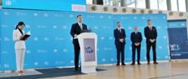 Na sali gimnastycznej z drewnianą podłogą stoi minister Czarnek za białą mównicą i mówi do mikrofonu, obok niego stoi kobieta w białym garniturze, z tyłu trzech mężczyzn w garniturach, za nimi niebieska ściana z napisem Program WF z AWF i duże okna.