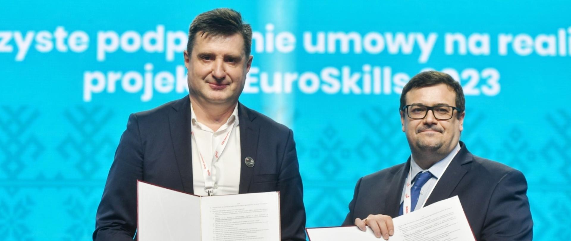 Dwóch mężczyzn w garniturach stoi na tle niebieskiej ściany, trzymają w rękach otwarte teczki z dokumentami.
