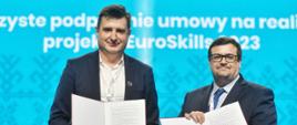 Dwóch mężczyzn w garniturach stoi na tle niebieskiej ściany, trzymają w rękach otwarte teczki z dokumentami.