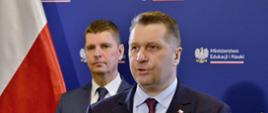 Minister Czarnek stoi i mówi do mikrofonu na stojaku, za nim wiceminister Piontkowski, za nimi niebieska ściana z napisem Ministerstwo Edukacji i Nauki, z boku polska flaga.