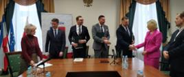 Przed ustawionymi w czworokąt drewnianymi stołami stoi pięciu mężczyzn w garniturach i dwie kobiety, jedna ubrana na czerwono stoi z boku, druga ubrana na fioletowo ściska rękę jednemu z mężczyzn, za nimi żółta ściana i dwa przysłonięte zasłonami okna.