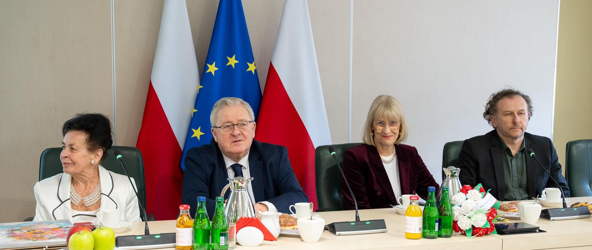 Stół za którym siedzi dwie kobiety i dwóch mężczyzn, za nimi flagi Polski i UE