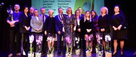 W ciemnej sali z błyszczącą podłogą stoi duża grupa ludzi, przed nimi stoją kwiaty, za nimi napis Najlepszy Nauczyciel Zawodu 2021 / 2022.