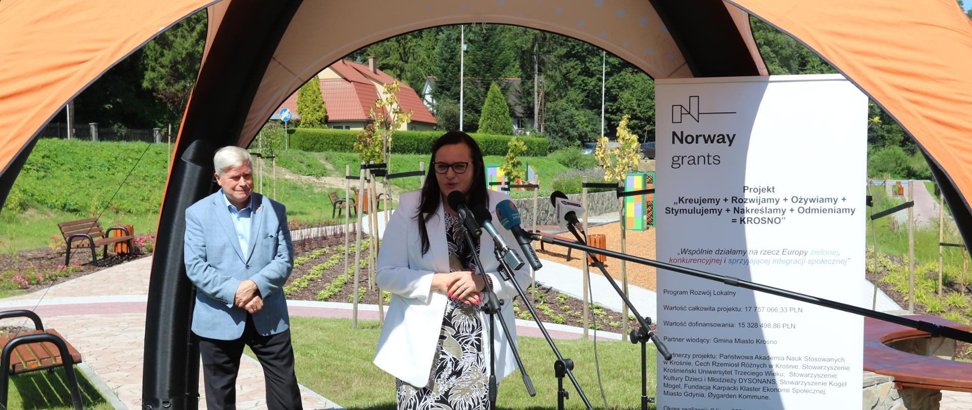 W parku pod namiotem przed mikrofonami stoi wiceminister Małgorzata Jarosińska-Jedynak. Za nią stoi mężczyzna.