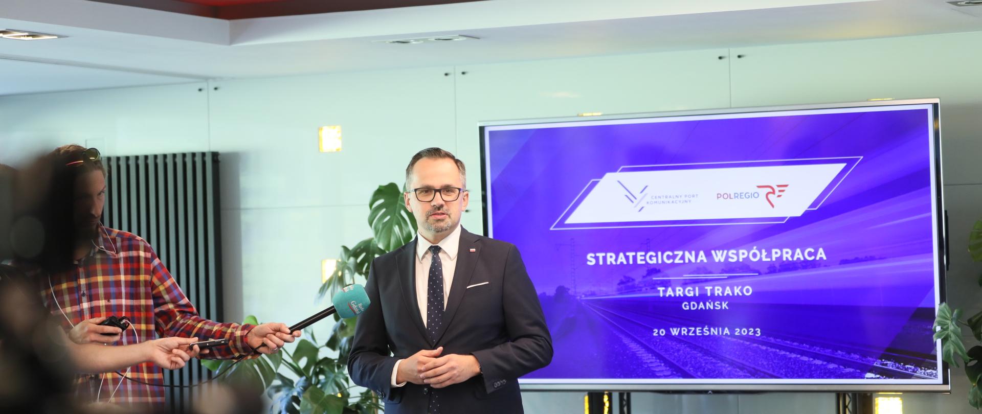 Wiceminister Marcin Horała stoi na sali i mówi do mikrofonu