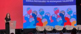 Przed zaciemnioną salą, w której w rzędach siedzą ludzie, stoi ciemno ubrana kobieta i mówi do mikrofonu, za nią kolorowa ściana z napisem Szkoła przyszłości to rozwijanie talentów.