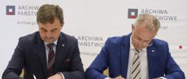 Dyrektor Adamiec i mężczyzna w czarnym garniturze siedzą za żółtym stołem, podpisują leżące przed nimi dokumenty.