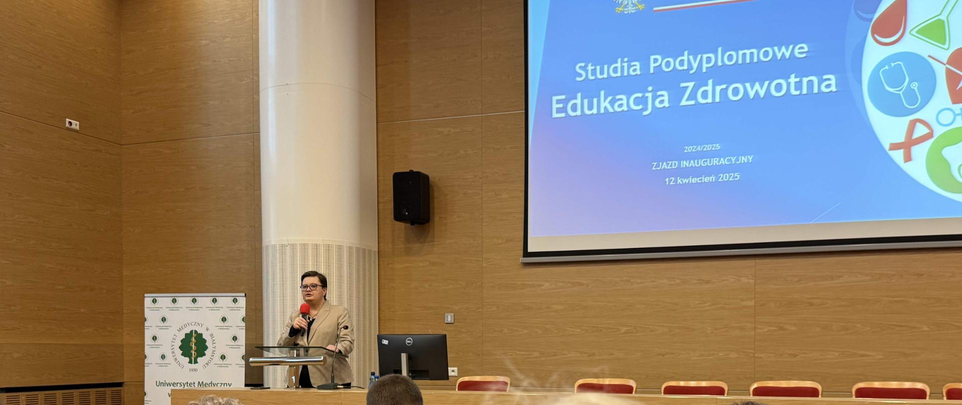 Widok z tyłu. Duża aula wykładowa. W ławach siedzą ludzie, przed nimi długi stół z krzesłami, po lewej stronie stoi kobieta przemawia do zebranych. Na ścianie duży ekran z napisem Studia podyplomowe edukacja zdrowotna.
