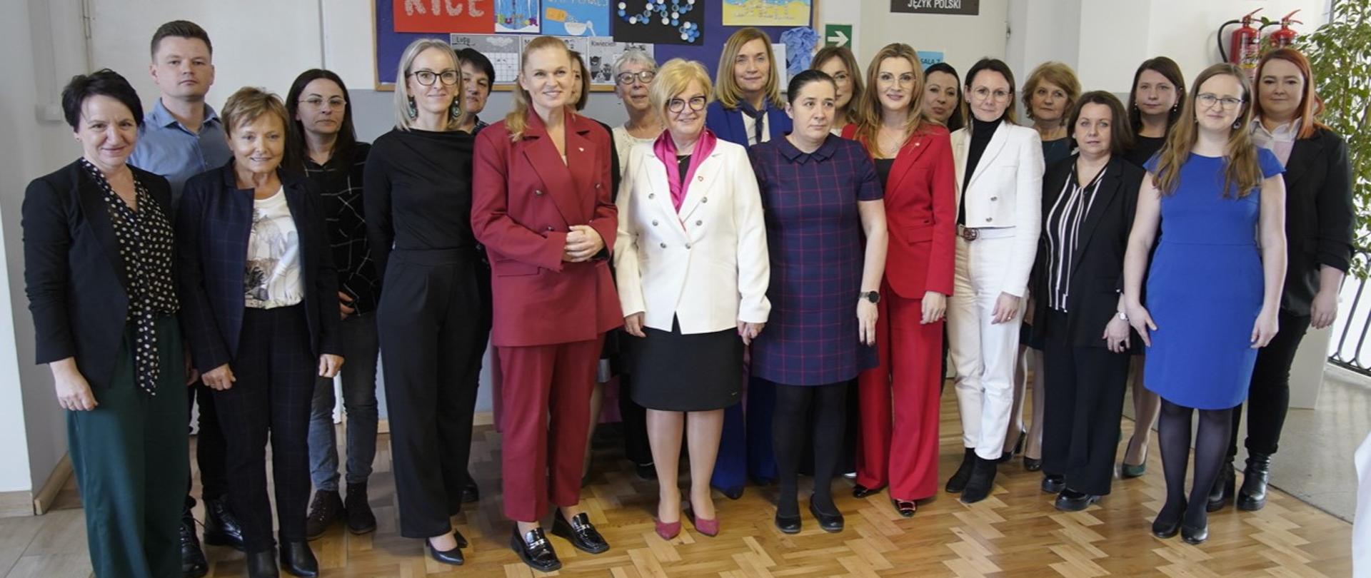 Minister Nowacka pozuje do zdjęcia w grupie kobiet i mężczyzn na korytarzu szkolnym. W tle widać tablice i drzwi do klas.