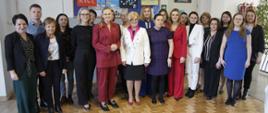 Minister Nowacka pozuje do zdjęcia w grupie kobiet i mężczyzn na korytarzu szkolnym. W tle widać tablice i drzwi do klas.